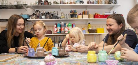 Kolme lasta istuu pöydän ääressä maalaamassa keskittyneen näköisenä. Mukana on myös kaksi hymyilevää aikuista.