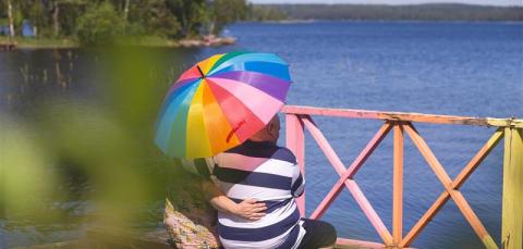 Kaksi henkilöä istuu laiturilla sateenkaarivärisen päivänvarjon alla kauniina kesäpäivänä.