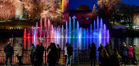 Värikkäitä vesisuihkuja Tanssivat vedet -näytöksessä Tammerkoskessa hämärän aikaan. Ihmiset seuraavat näytöstä kaiteen äärellä.