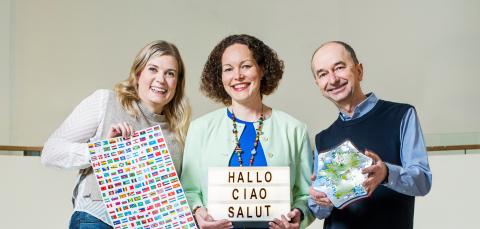 Kolme henkilöä seisoo vierekkäin. Heillä on käsissään kielten opiskelutavaroita, kuten lippuja ja taulu.