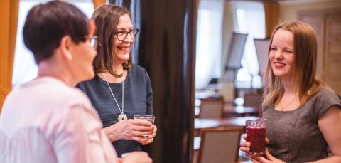 Kolme hymyilevää naista seisoo juomalasit kädessään keskustelemassa.