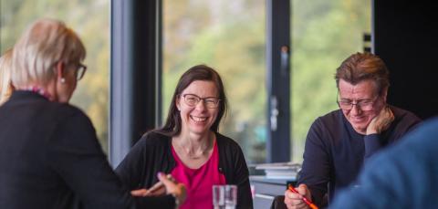 Henkilöitä istuu pöydän ääressä ja hymyilee.