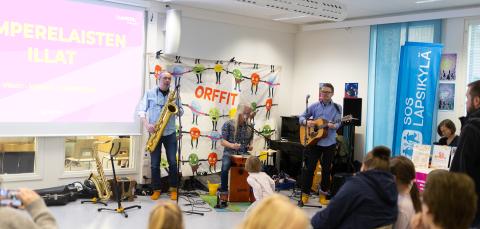 Lastenmusiikkiyhtye soittaa koulun tiloissa. Yleisössä on eri-ikäisiä ihmisiä.