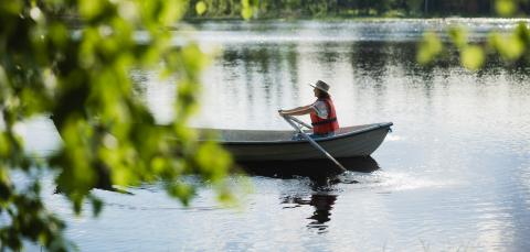 Henkilö soutaa soutuveneellä kesäisellä järvellä olkihattu päässä ja päällään pelastusliivit.