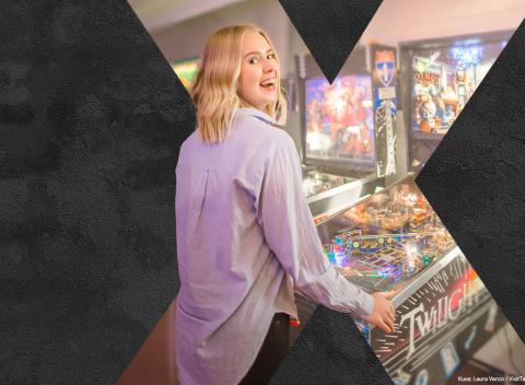 Mustalla taustalla on vaaleahiuksinen hymyilevä nainen, joka pelaa flipperillä.