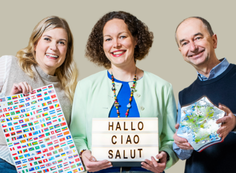 Kolme henkilöä seisoo vierekkäin. Heillä on käsissään kielten opiskelutavaroita, kuten lippuja ja taulu.