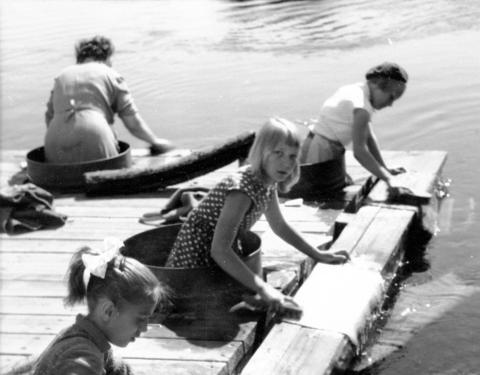 Lapsia pesemässä mattoja. Vaaleatukkainen tyttö katsoo kameraan.