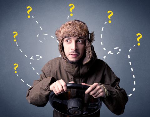 Nuori mies seisoo harmaata seinää vasten ja pitelee ohjauspyörää. Seinällä menee polkuja, joiden päässä on kysymysmerkkejä. Miehellä on karvalakki, ja hän katsoo silmät pyöreinä sivulle.