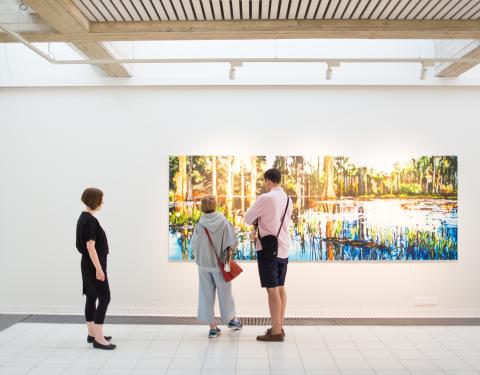 Kolme henkilöä katselee isoa värikästä maisemataulua.