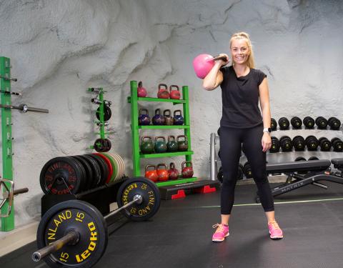 Henkilö pitelee isoa kahvakuulaa yhdellä kädellä käsi koukussa luolamaisessa kuntosalissa.