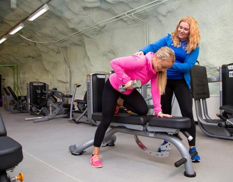 Ohjaaja ohjaa asentoa. Treenaaja nostaa käsipainoa penkillä.