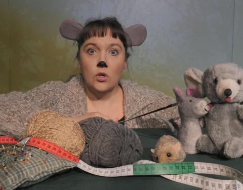 Hiireksi naamioitunut nainen, joka kertoo satua erilaisten pehmolelujen kera.