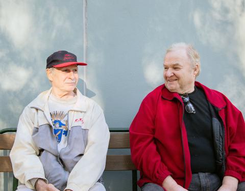 Kaksi miestä istuu ulkona seinän viereen asetetulla penkillä.