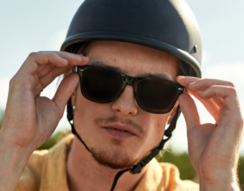 Nuori mies seisoo pihalla kypärä ja aurinkolasi päässään. Hän pitelee aurinkolaseja niiden sangoista.