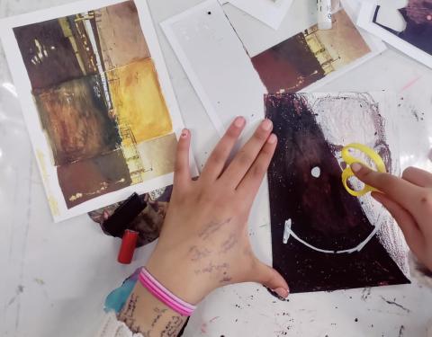 Kuvassa pöytä, kuvataidetarvikkeita, papereita ja oman teoksensa parissa työskentelevän nuoren kädet. Nuoren teos on mustavalkoinen ja siinä on yksinkertaiset hymyilevät kasvot.