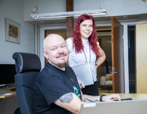 Kaksi kohti katsovaa hymyilevää henkilöä työpöydän ääressä. Toinen istuu tietokoneella, toinen seisoo taustalla.