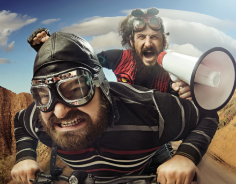 Mies ajaa polkupyörällä. Hänellä on ajolasit ja hän nojaa eteenpäin. Hänen tarakallaan istuu toinen mies, joka huutaa megafoniin ja nostaa kättään ylös.