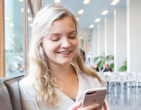 Nuori nainen istuu isossa ja avarassa tilassa sohvalla. Hän katsoo kännykkäänsä ja hymyilee.