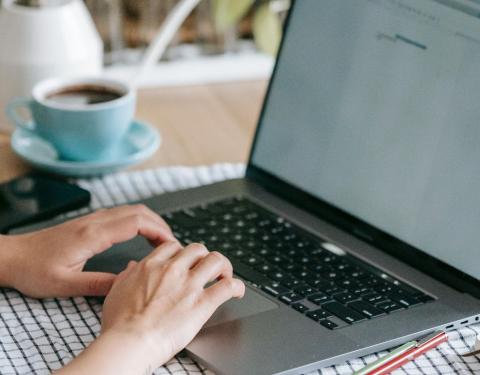 Pöydällä on kahvikuppi ja tietokone. Kädet ovat näppäimistöllä.