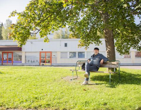 Vaalea matala talo. Sen edustalla puu ja puun alla penkillä ihminen istumassa.