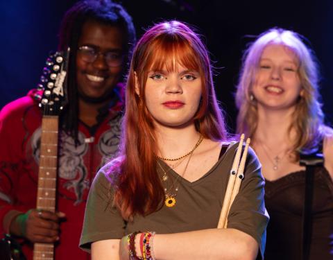 Kolme nuorta, yhdellä on rumpukapulat, joissa on silmät. Toisella on kitara kädessään.