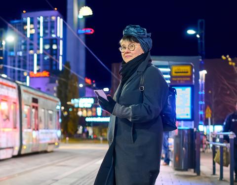 Henkilö seisoo ratikkapysäkillä ilta-aikaan, kädessä älypuhelin.