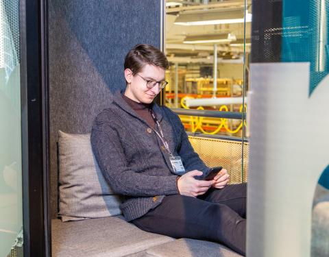 Henkilö istumassa puhelinkopissa ja katsoo hymyillen sylissään olevaa kännykkää.