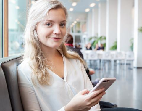 Nuori hymyilevä opiskelija istuu yliopiston käytävällä ja selaa kännykkäänsä.