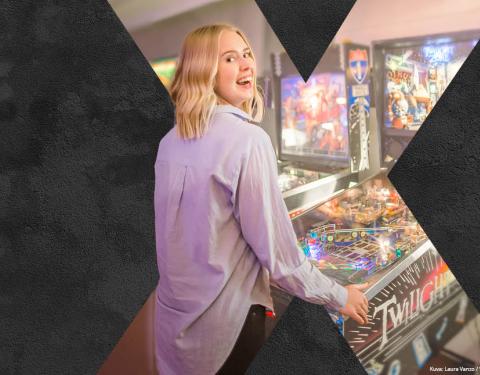 Mustalla taustalla on vaaleahiuksinen hymyilevä nainen, joka pelaa flipperillä.