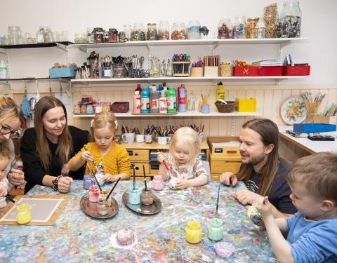 Kolme aikuista ja neljä pikkulasta ison pöydän ääressä. Lapset tutkivat keskittyneen näköisenä värikkäitä maalipurkkeja. Taustalla näkyy hyllykkö, jossa on siistissä järjestyksessä erilaisia kuvataidetarvikkeita.