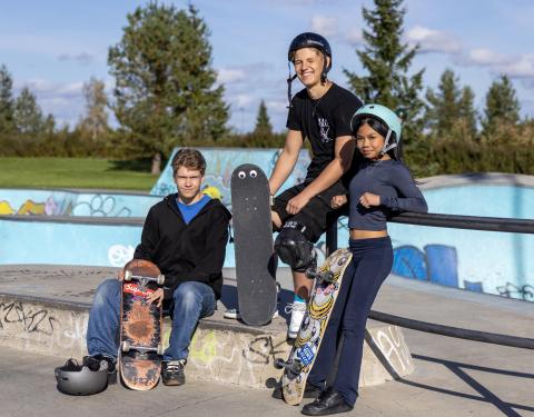 Kolme henkilöä katsoo kameraan skeittilautojen kanssa.