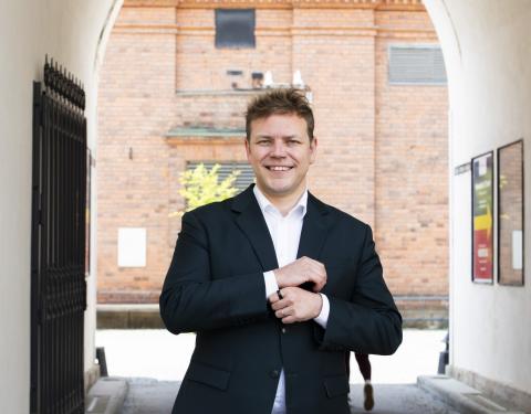 Henkilö seisoo kädet edessään ulkona valkoisten seinien välissä takanaan tiiliseinää.