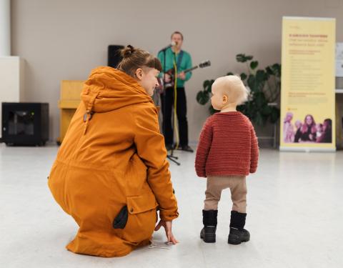 Oranssitakkinen äiti on kyykyssä pienen lapsensa vieressä. He katselevat toisiaan. Taustalla näkyy muusikko.