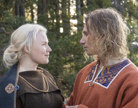 Muinaispukuihin pukeutuneet nainen ja mies katsovat toisiaan kesäisen metsämaiseman edessä.