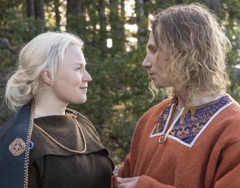 Muinaispukuihin pukeutuneet nainen ja mies katsovat toisiaan kesäisen metsämaiseman edessä.