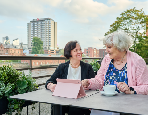Kaksi ikääntynyttä henkilöä istuu pöydän ääressä edessään tablet-laite ja kahvikuppi.