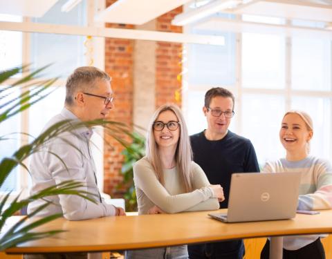 Four smiling people are standing at a desk in an office, chatting. There is a computer on the desk.