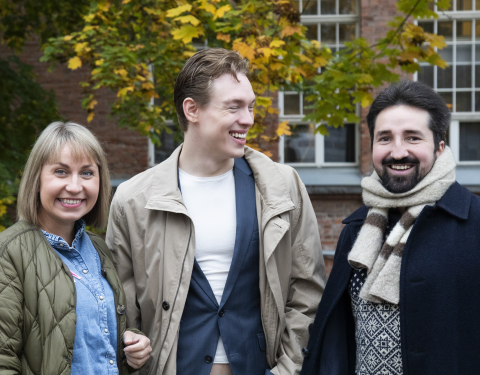 Kolme henkilöä seisoo syksyisen värikkään puun edessä.