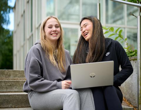 Kaksi henkilöä istuu hymyillen portailla. Toisella on kannettava tietokone sylissään.
