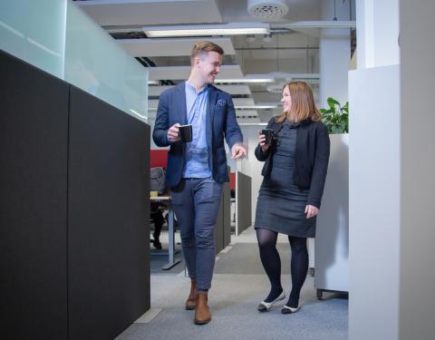 Kaksi henkilöä kävelee kahvikupit kädessä toimistorakennuksessa.