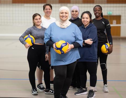 Six women on a volleyball court in a group photo. All of them smiling for a camera