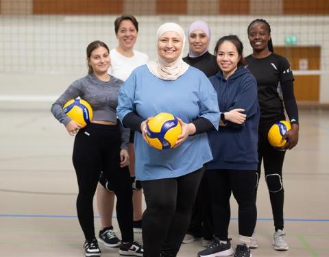 Six women on a volleyball court in a group photo, three of them are holding a volleyball