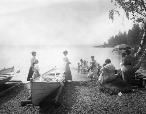 Vanha valokuva, jossa henkilöt ovat järvenrannalla. Edessä veneitä ja reunalla puita.