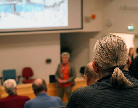 Henkilöt istuvat luentosalissa. Taustalla on valkokangas ja luennoitsija.