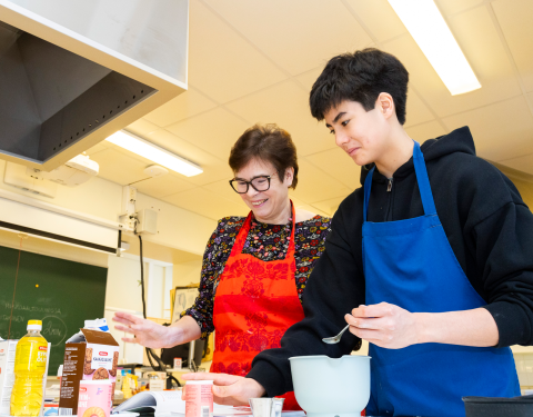 Kaksi henkilöä on leivontatarvikkeiden ääressä esiliinat edessään.