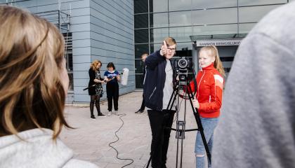 Kaksi opiskelijaa kameran takana, toinen näyttää sormellaan ylöspäin. Etualalla kuvattavia.