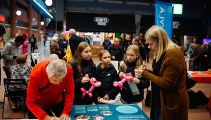 People of different ages at the Tampere Residents’ Evening in a shopping center. At the table, there are two adults writing on paper and three young people with balloons.