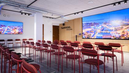 Kuvassa on Siluetin esitystila. Kuvan etualalla on neljä riviä punaisia penkkejä ja takana on vaalea, puinen seinä jolla on suuri näyttö. Näytössä on kuva Tampereen keskustasta.