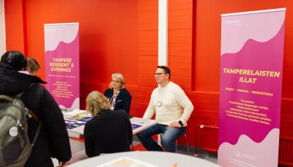 Kaksi kaupungin työntekijää istuu pöydän takana tamperelaisten illassa. Heidän takanaan on voimakkaan oranssi seinä.