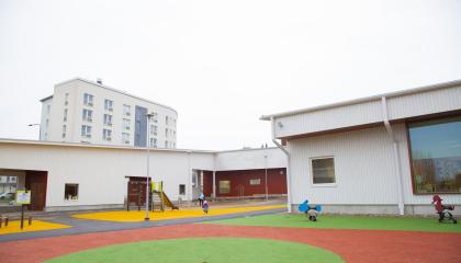 Hippoksen valkoisen puupäiväkodin pihalla näkyy värikäs pelialue.